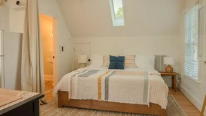 Camera da letto con soffitto spiovente, un letto grande con copriletto bianco a righe, due lampade da comodino e una finestra con persiane.