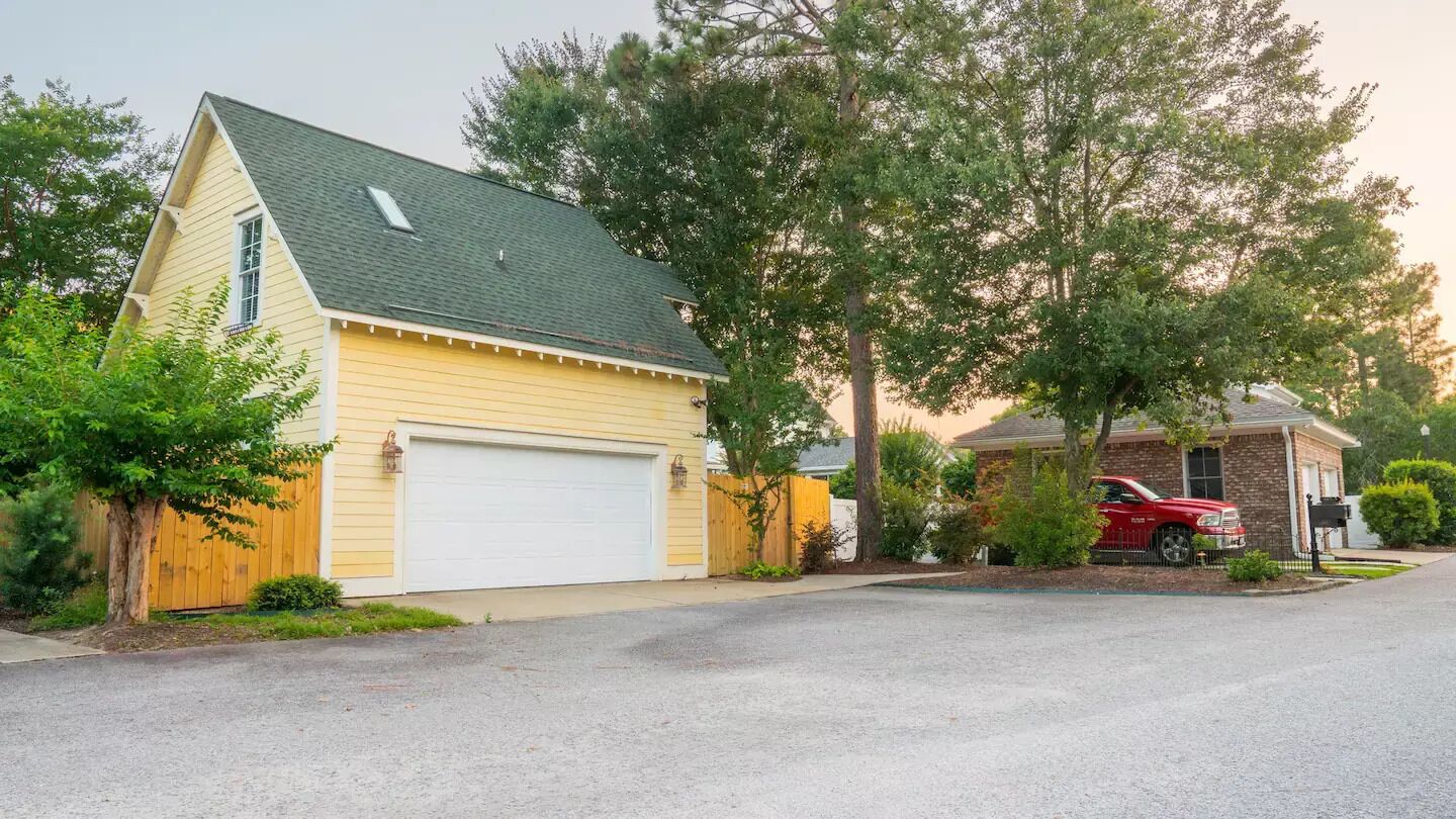 Een woonhuis met een garage, een rode auto ervoor geparkeerd, en een grote boom aan de rechterkant.