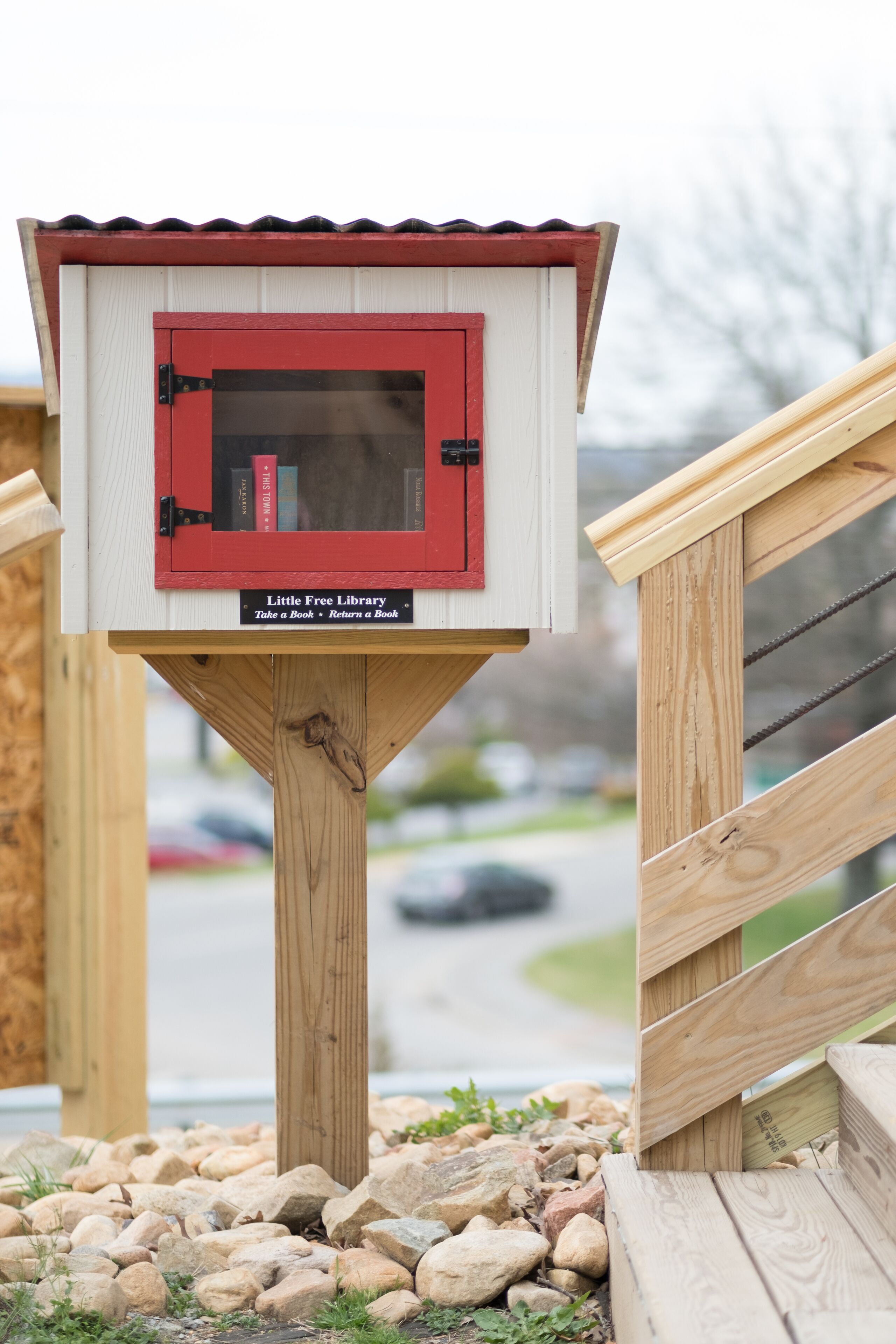 Eine kleine Holzhütte mit roter Tür, beschriftet als „Little Free Library“, mit einem Schild darunter: „Buch mitnehmen – Buch zurückgeben.“