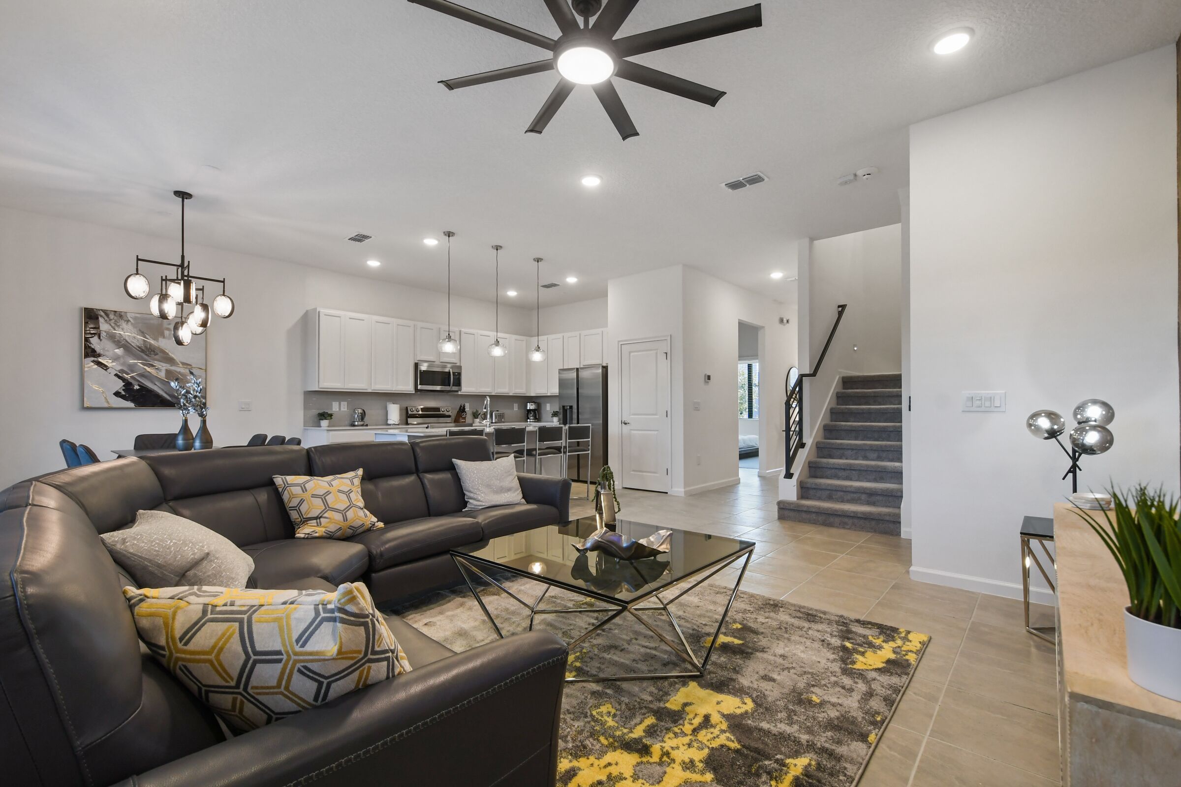 A modern living room with a black leather sofa, glass coffee table, and a contemporary chandelier. A kitchen is visible in the background with white cabinets and stainless steel appliances.