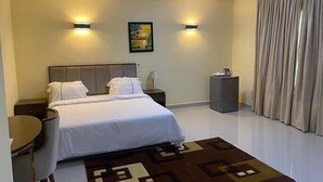 A hotel room with a bed, bedside tables, a desk, a chair, a lamp, a painting on the wall, and a window with curtains. - Premium Hotel & Suites by Victoria Inn (Benin City)