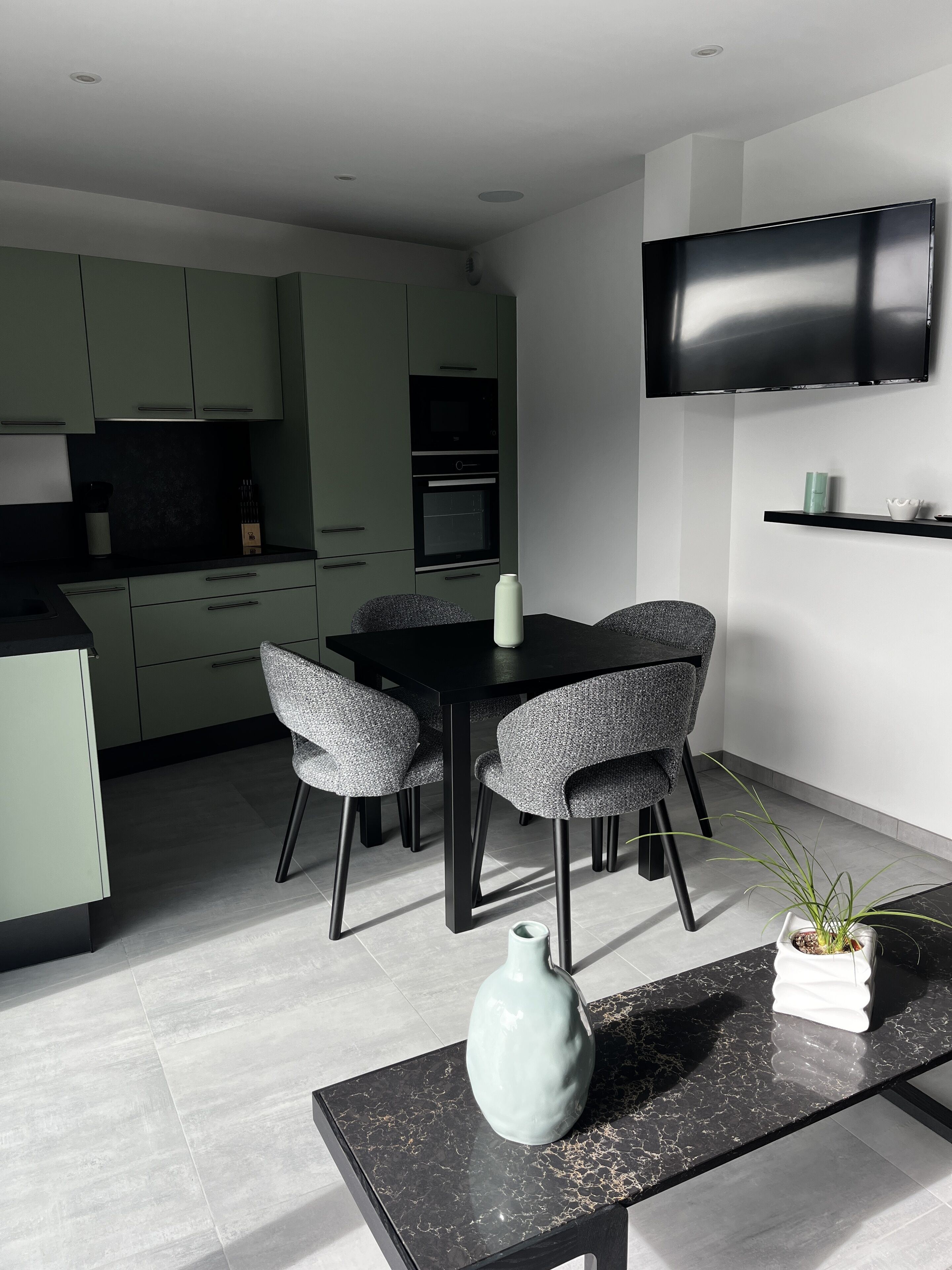 Une cuisine moderne avec un coin repas, comprenant une table noire, des chaises à revêtement gris, un vase blanc et un téléviseur à écran plat fixé au mur.