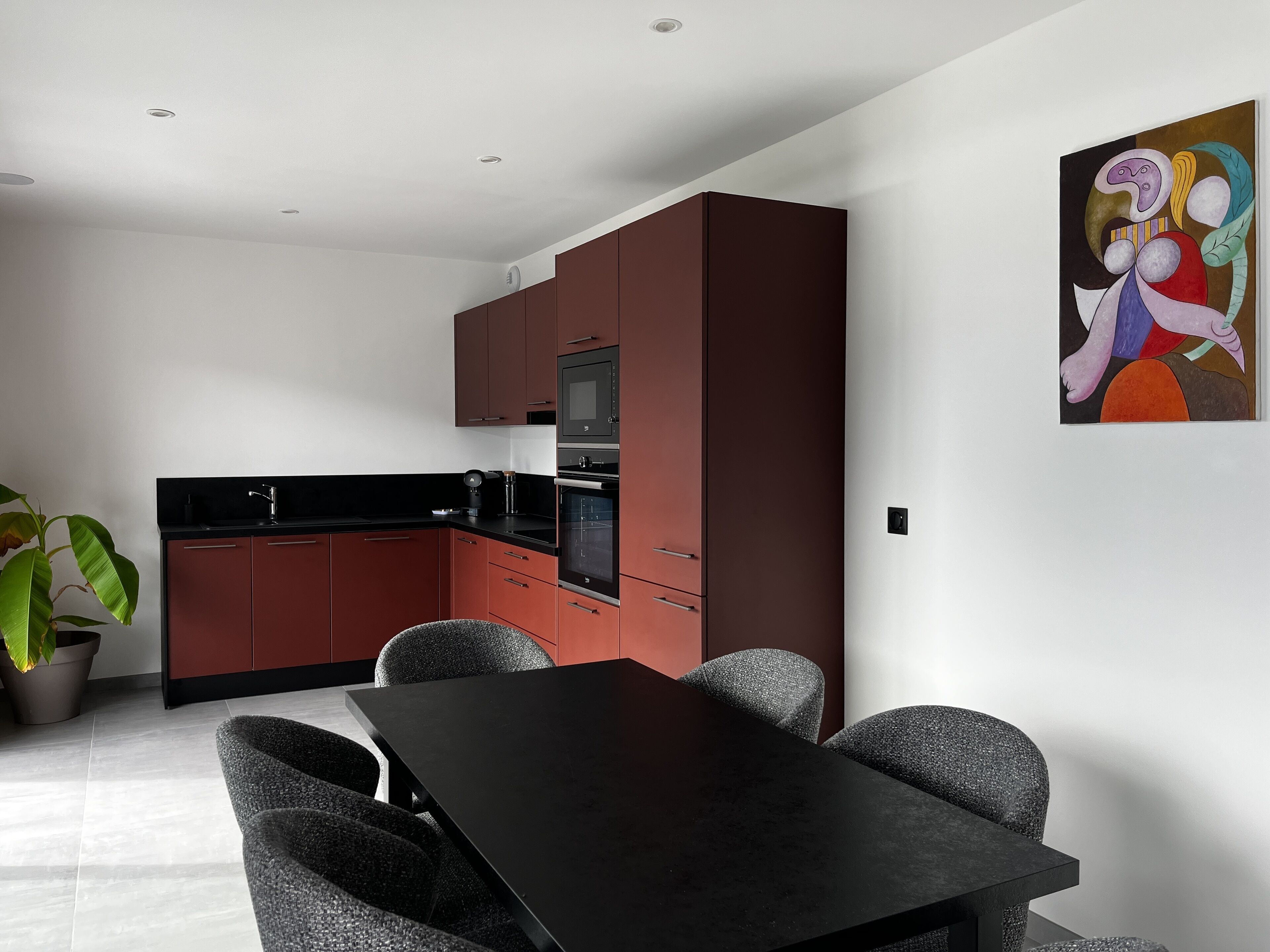 A modern kitchen with dark red cabinets, a black countertop, and a stainless steel oven. There is a dining table with four chairs.