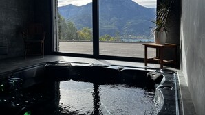 A room with a large window overlooking mountains, a hot tub, a chair, and a potted plant. - Les Restanques du Lac (Savines-le-Lac)
