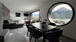 A modern living room with a dining area, a large round window offering a mountain view, and a flat-screen TV mounted on the wall. - Les Restanques du Lac (Savines-le-Lac)