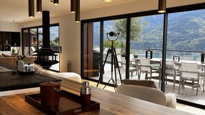 A modern kitchen with a wooden countertop, a coffee machine, and a kettle. There is a large window with a view of mountains and a balcony with outdoor seating. - Les Restanques du Lac (Savines-le-Lac)