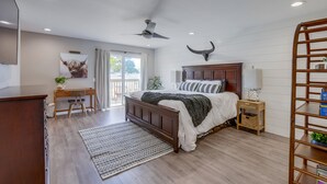 Un dormitorio con una cama de madera, un escritorio, una silla, un ventilador de techo y un ventanal grande.