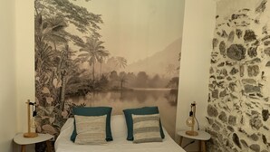A bedroom with a large mural of a tropical landscape, a white bed with two pillows, wooden flooring, and a stone wall on one side. - Gîte Dans les Cévennes (L'Estréchure)