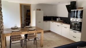 A compact kitchen with white cabinets, a black oven, and a dining area with a wooden table and wicker chairs. - Gîte Dans les Cévennes (L'Estréchure)
