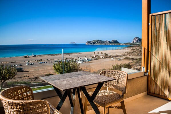 A balcony with wicker chairs and a table overlooking a beach with boats and a clear sky. - Panorama Hotel (Kissamos)