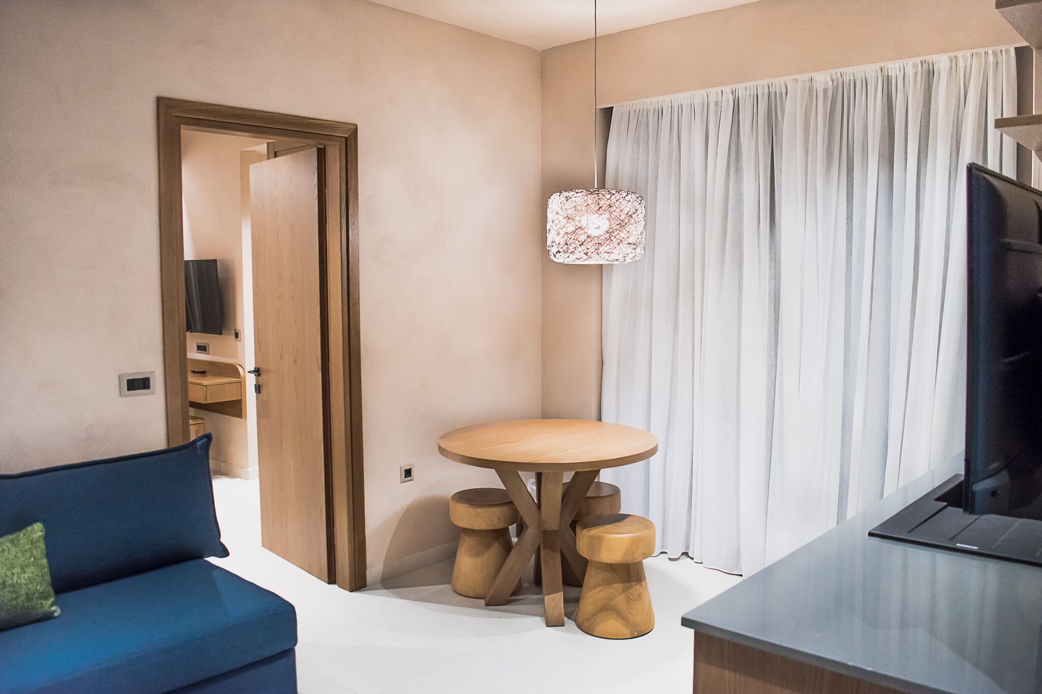 Una habitación de hotel moderna con una mesa redonda de madera, un sofá azul, un televisor y un ventanal con cortinas.
