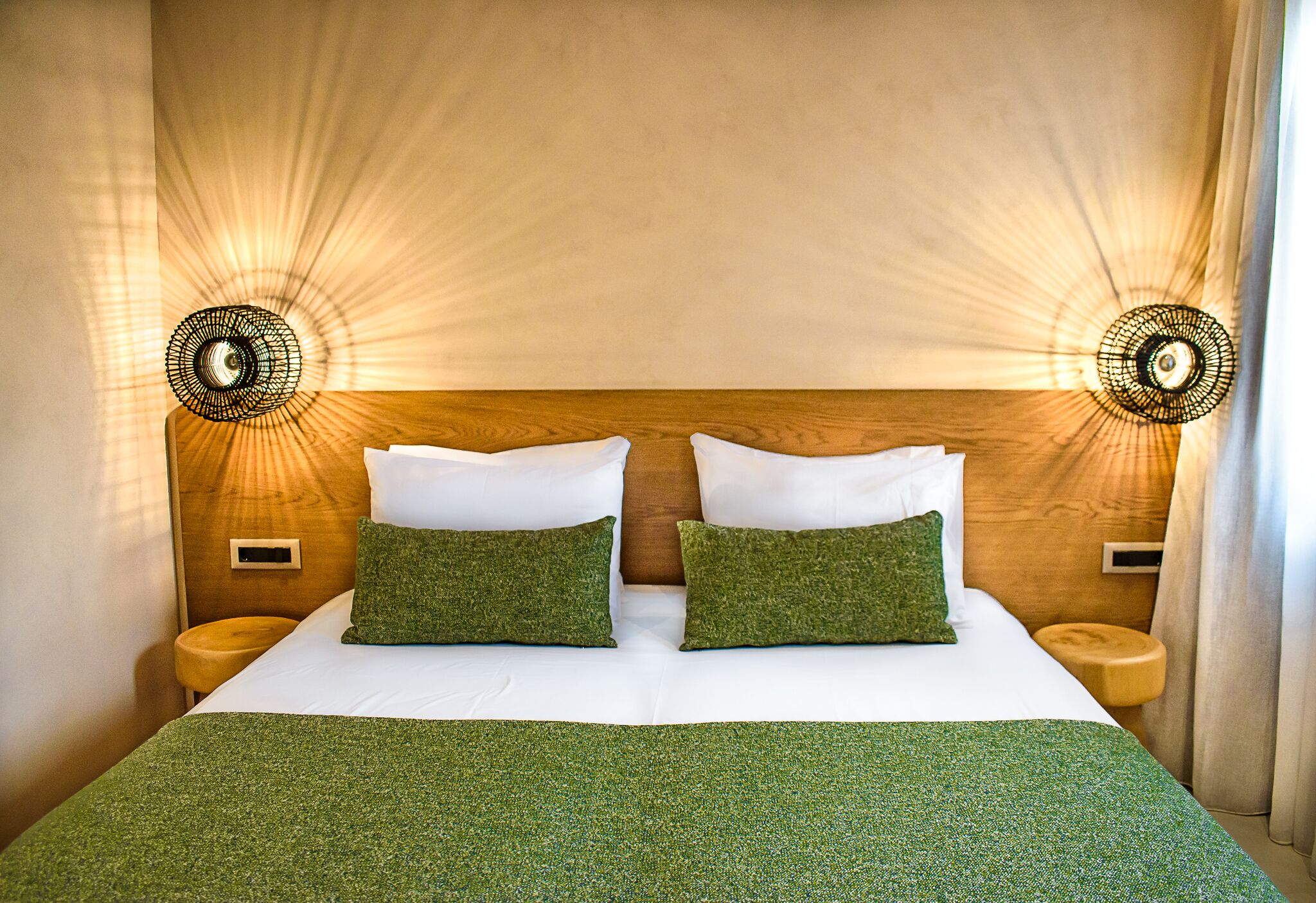 Una cama bien tendida con una colcha verde y dos almohadas, con dos lámparas de pared de diseño circular a cada lado.