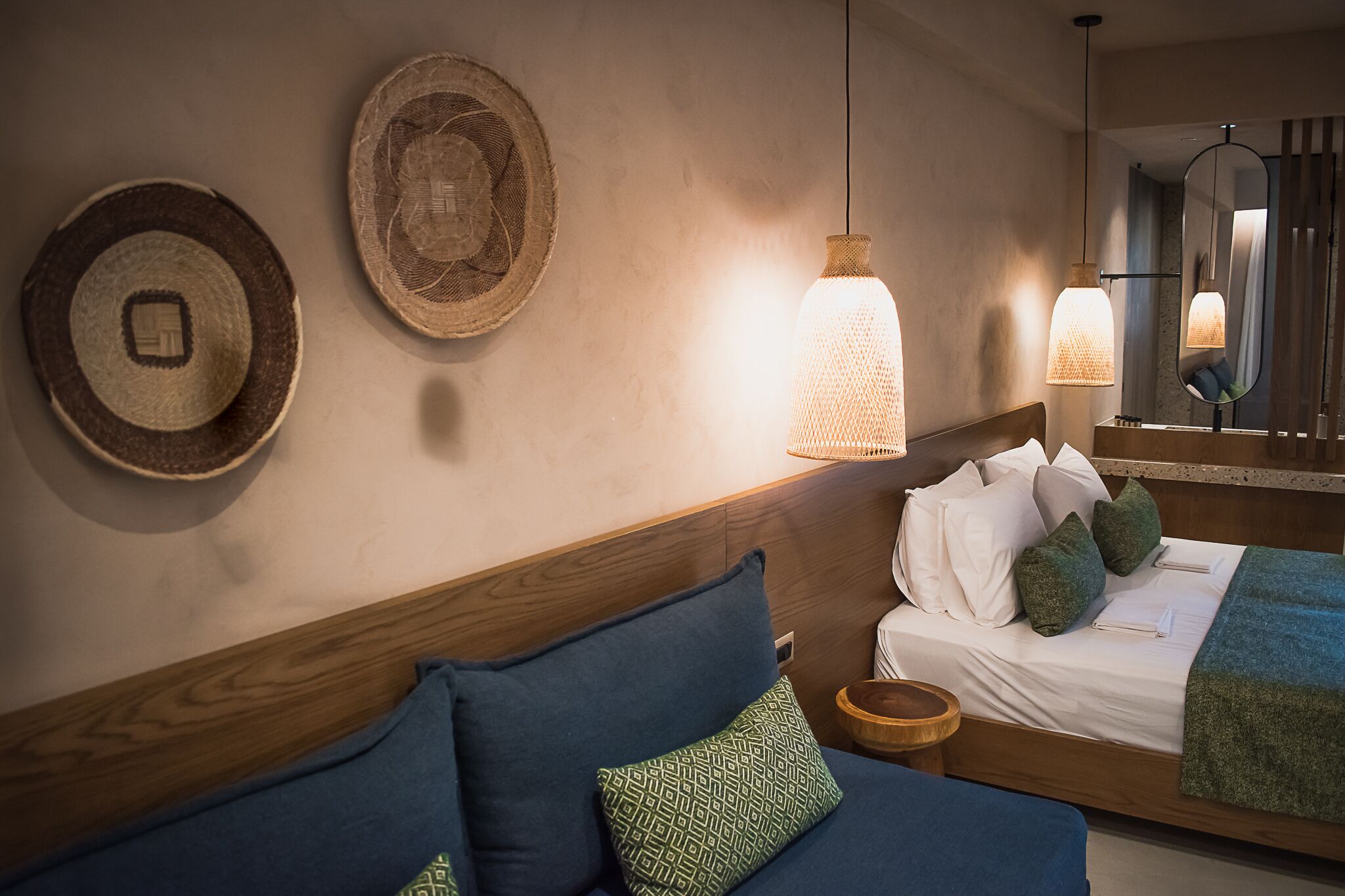 A hotel room with a wooden headboard, a bed with white linens and green pillows, a round woven wall hanging, and a hanging pendant light.