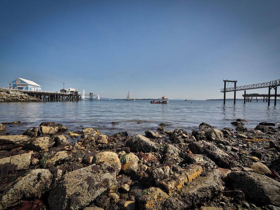 A rocky shoreline with a pier extending into calm waters, a small boat, and a distant sailboat.