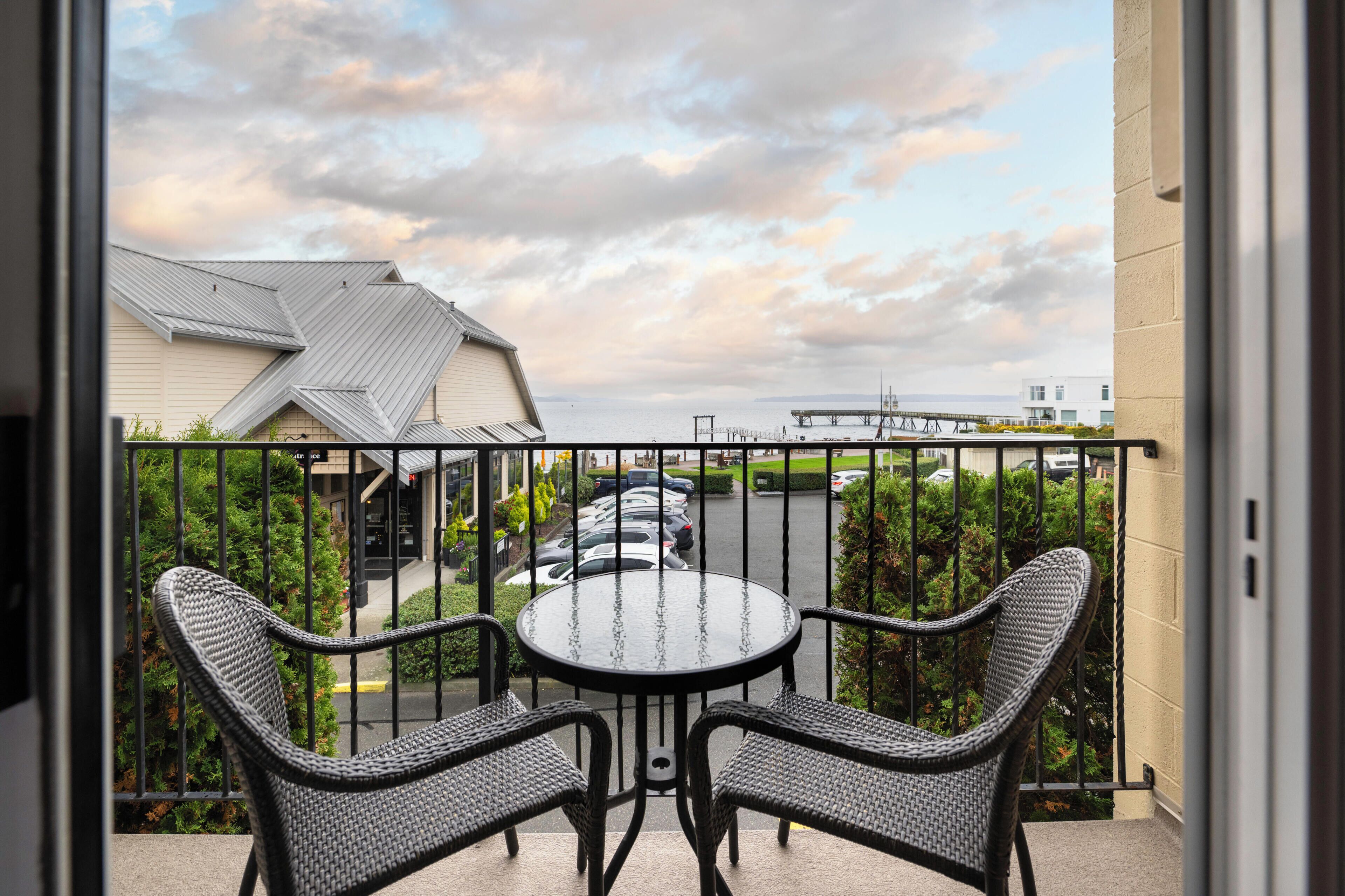 Een balkon met twee rieten stoelen en een klein rond tafeltje, met uitzicht op een parkeerterrein en gebouwen.