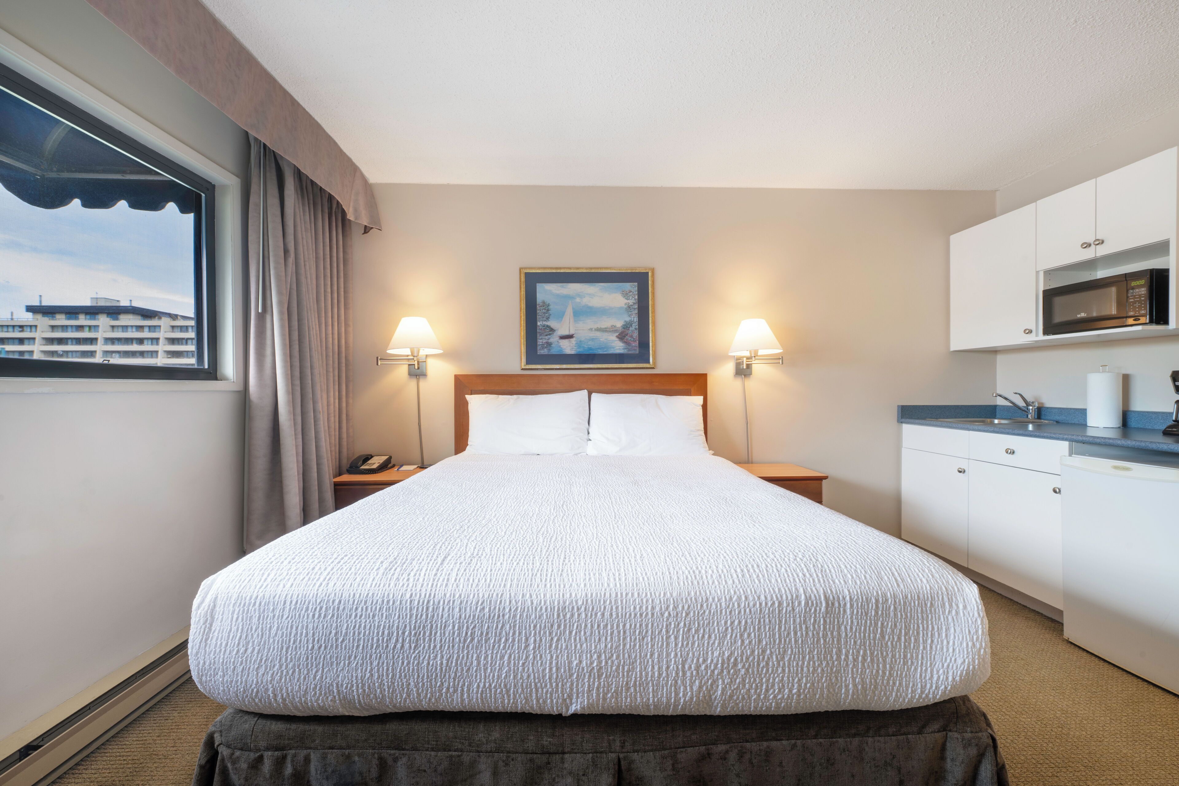 A hotel room with a large bed, two bedside lamps, a painting on the wall, a window with curtains, a kitchenette with a microwave, and a view of a building outside.