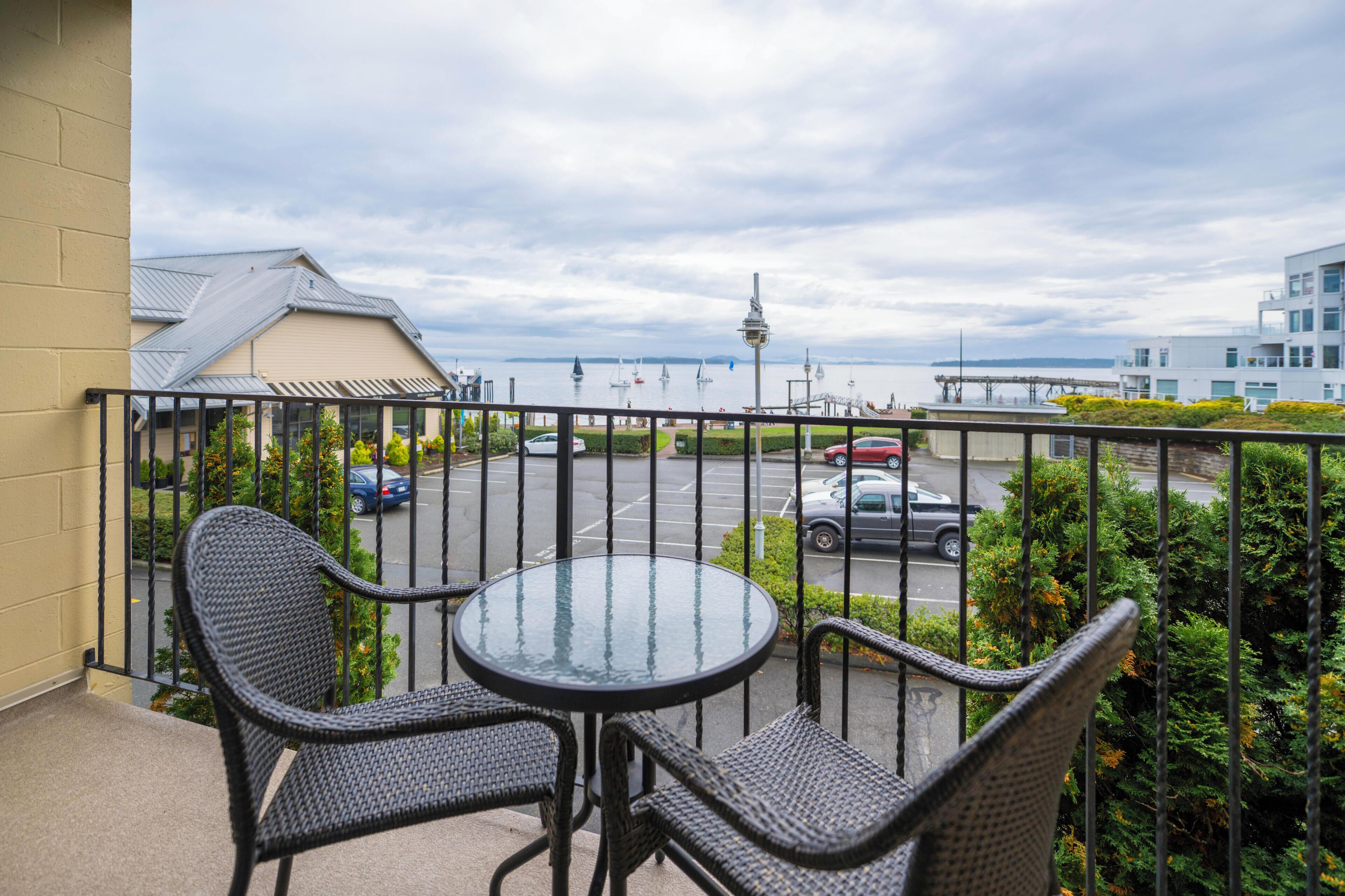 A balcony with wicker chairs and a glass table, overlooking a parking area and a distant view of a cityscape with a tower.