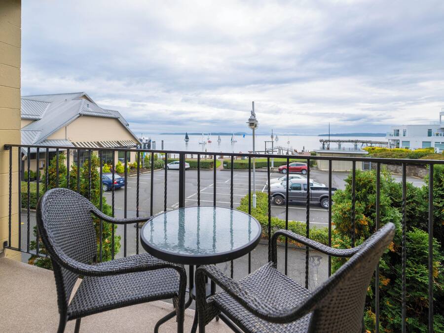 A balcony with wicker chairs and a glass table, overlooking a parking area and a distant view of a cityscape with a tower.