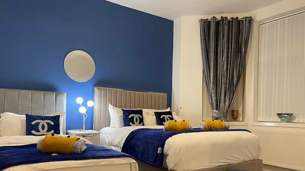 A hotel room with two beds, a round mirror, a nightstand with a lamp, and a window with curtains. - COSY HLA HOLYTOWN APARTMENT (Bellshill)