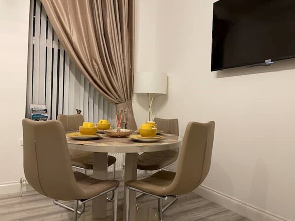 A dining area with a round table, four chairs, a flat-screen TV mounted on the wall, and a window with vertical blinds. - COSY HLA HOLYTOWN APARTMENT (Bellshill)
