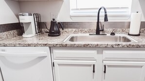 Una cocina con armarios blancos, encimera de granito, cafetera negra, hervidor eléctrico negro, pileta de acero inoxidable y lavavajillas blanco.