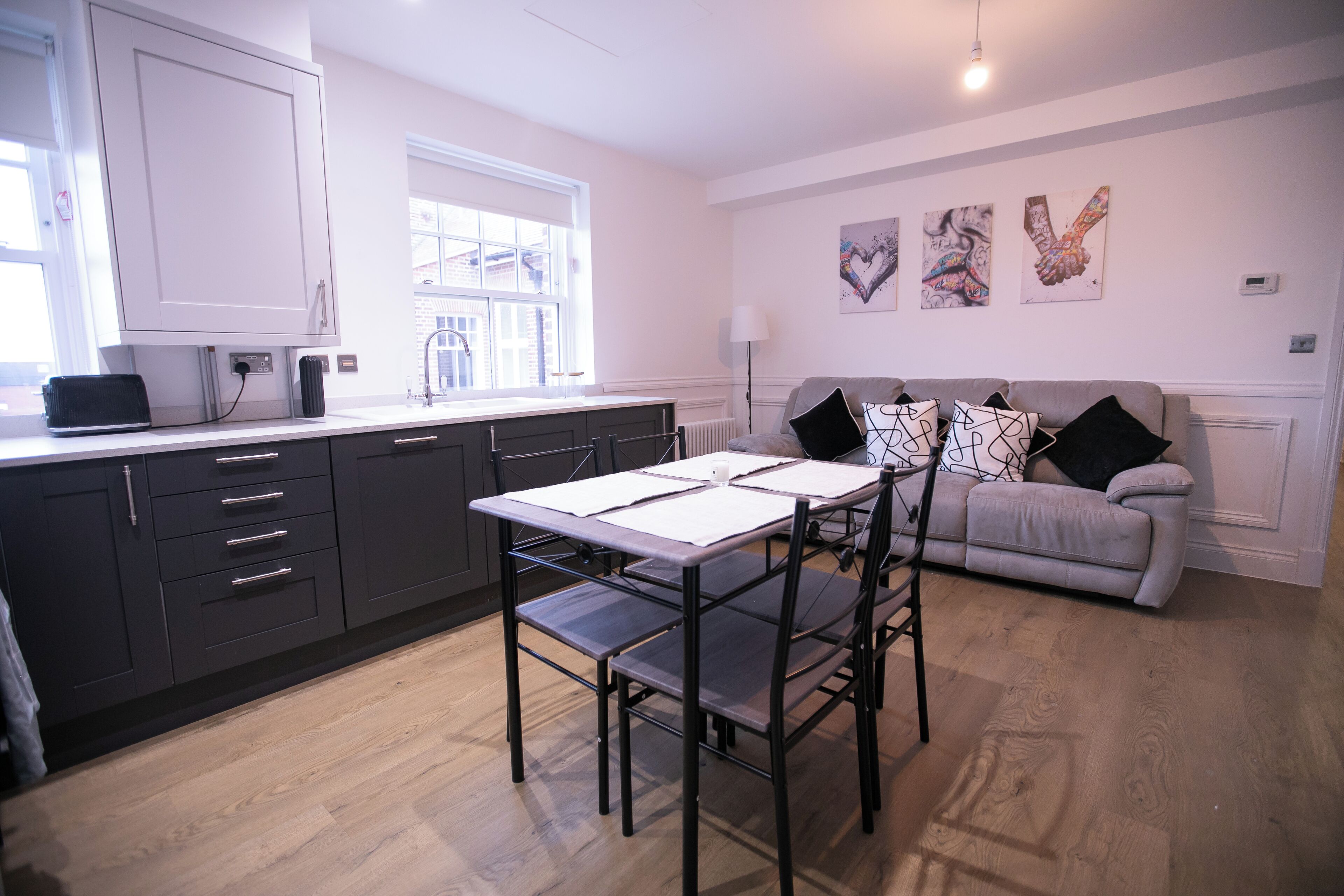 A modern kitchen with dark cabinets, a white countertop, and a dining table with chairs. There is a grey sofa with black cushions in the living area, and artwork on the wall.