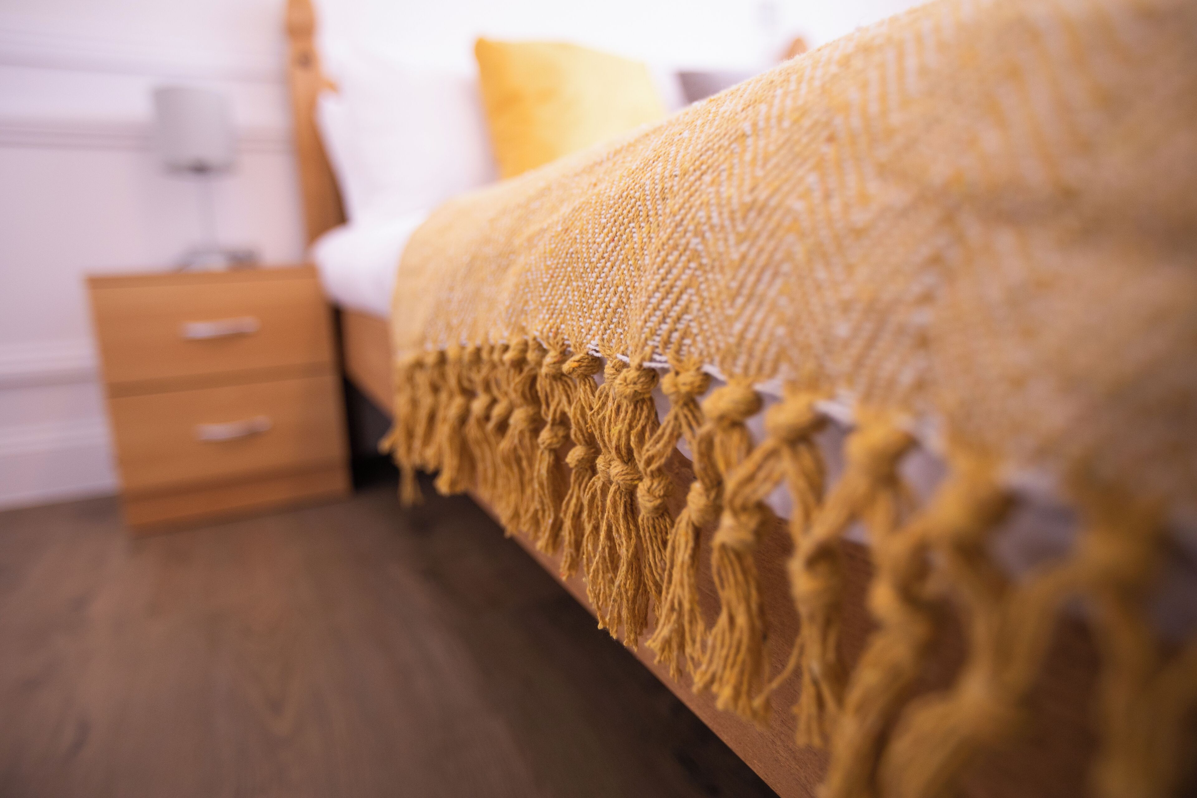 Una cama con un edredón texturizado y flecos, una mesita de noche de madera con cajones y un almohadón amarillo.