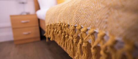 Una cama con un edredón texturizado y flecos, una mesita de noche de madera con cajones y un almohadón amarillo.