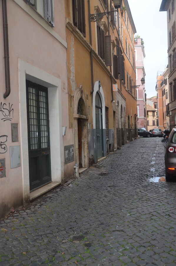 A narrow cobblestone street with historic buildings, a closed shop with a barred window, and parked cars.