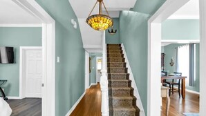 A hallway with a wooden floor, a staircase with patterned carpet, and a chandelier.
