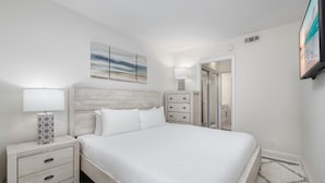 Un dormitorio con cabecera de madera, ropa de cama blanca, una mesita de noche con lámpara, cómoda y televisor montado en la pared.