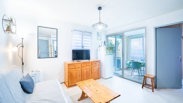 A bright, modern room with a bed, wooden bench, a television on a cabinet, a mirror, and a sliding glass door leading to a balcony.