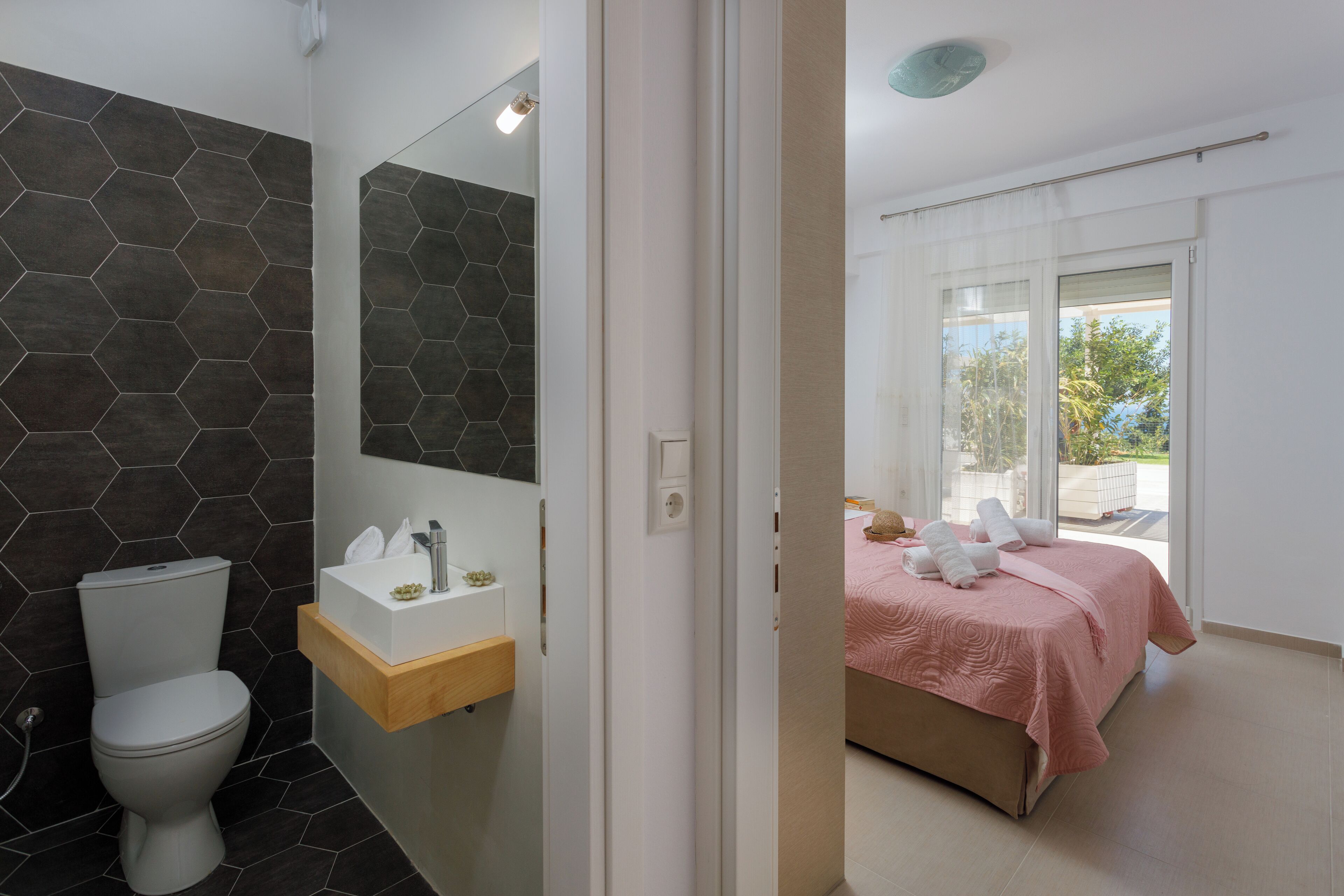 Une salle de bain moderne avec un mur recouvert de carreaux hexagonaux, des toilettes, un lavabo doté d’un plan de travail en bois et un miroir.