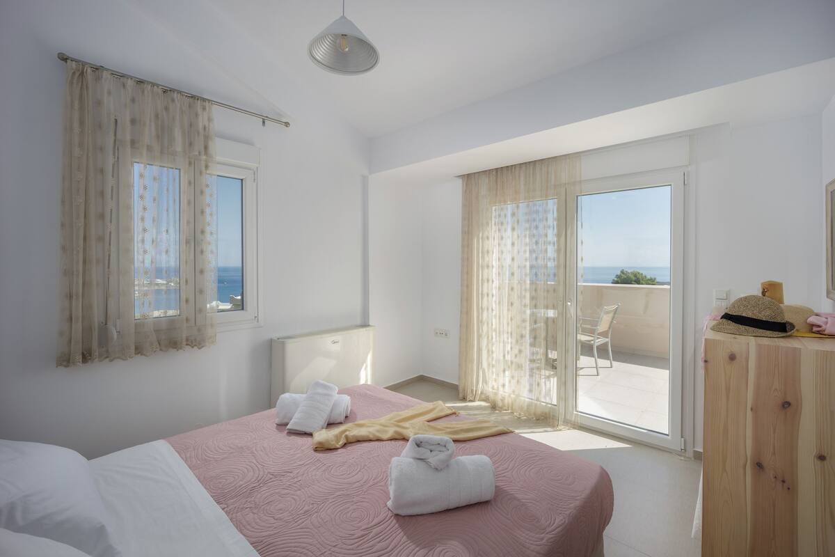 A bedroom with a bed, a wooden dresser, a balcony with a chair, and a view of the sea.