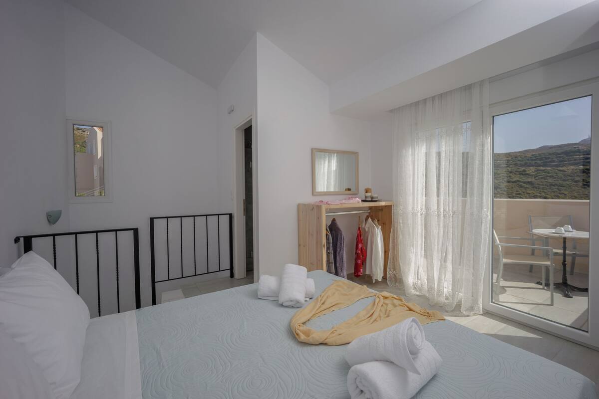 A hotel room with a bed, a vanity with a mirror, a balcony with a table and chairs, and a view of the landscape.