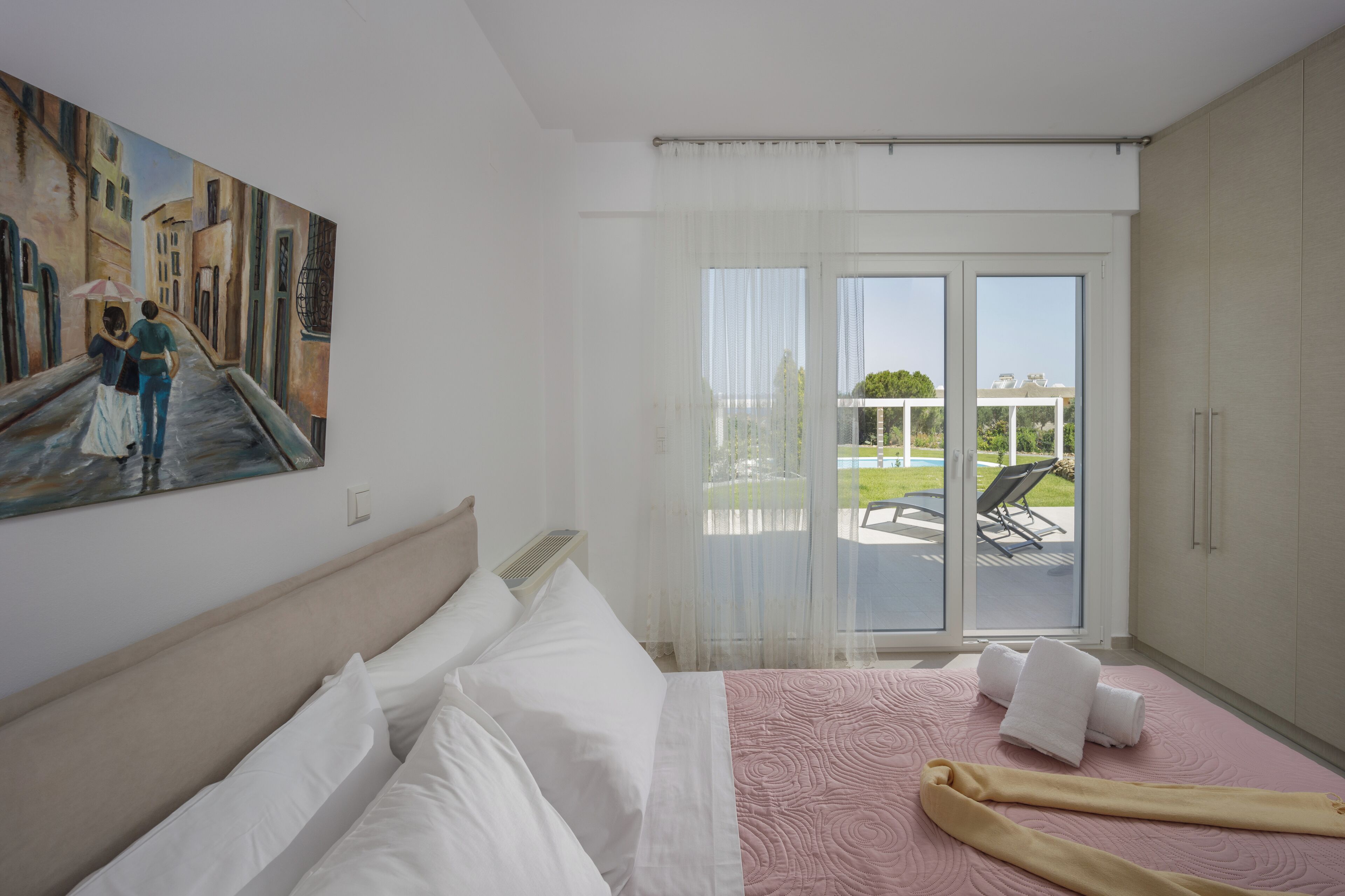 A hotel room with a bed, a painting on the wall, and a balcony with a view of the outdoors.