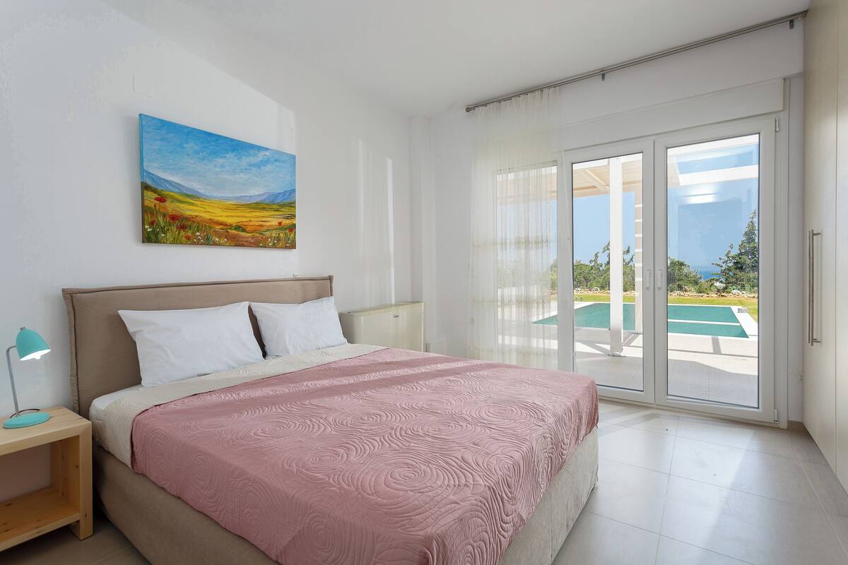 A bedroom with a bed, a nightstand, a painting, and a view of an outdoor swimming pool through a sliding glass door.