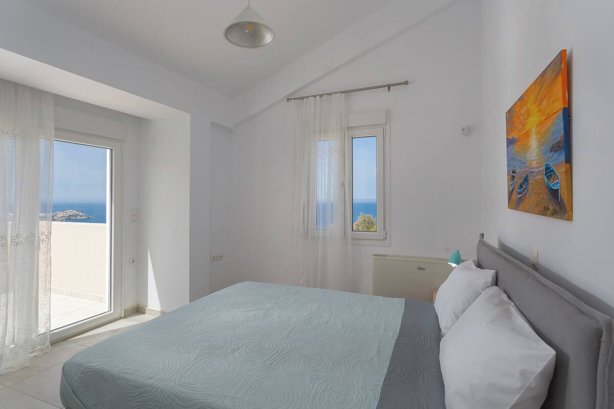 A bedroom with a bed, a window with curtains, a painting on the wall, and a view of the sea.