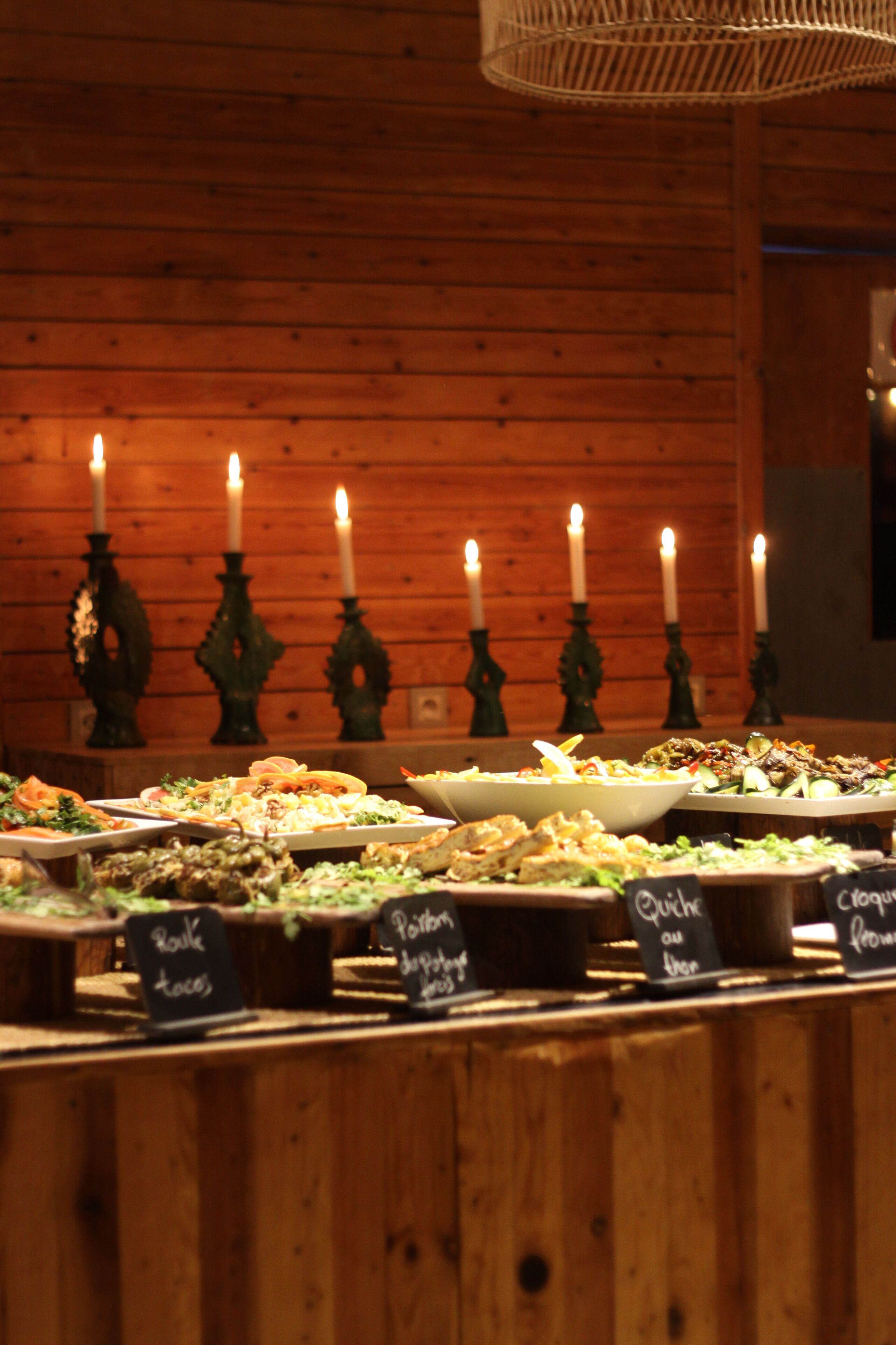 Buffet com diversos pratos identificados, velas ao fundo e painéis de madeira.