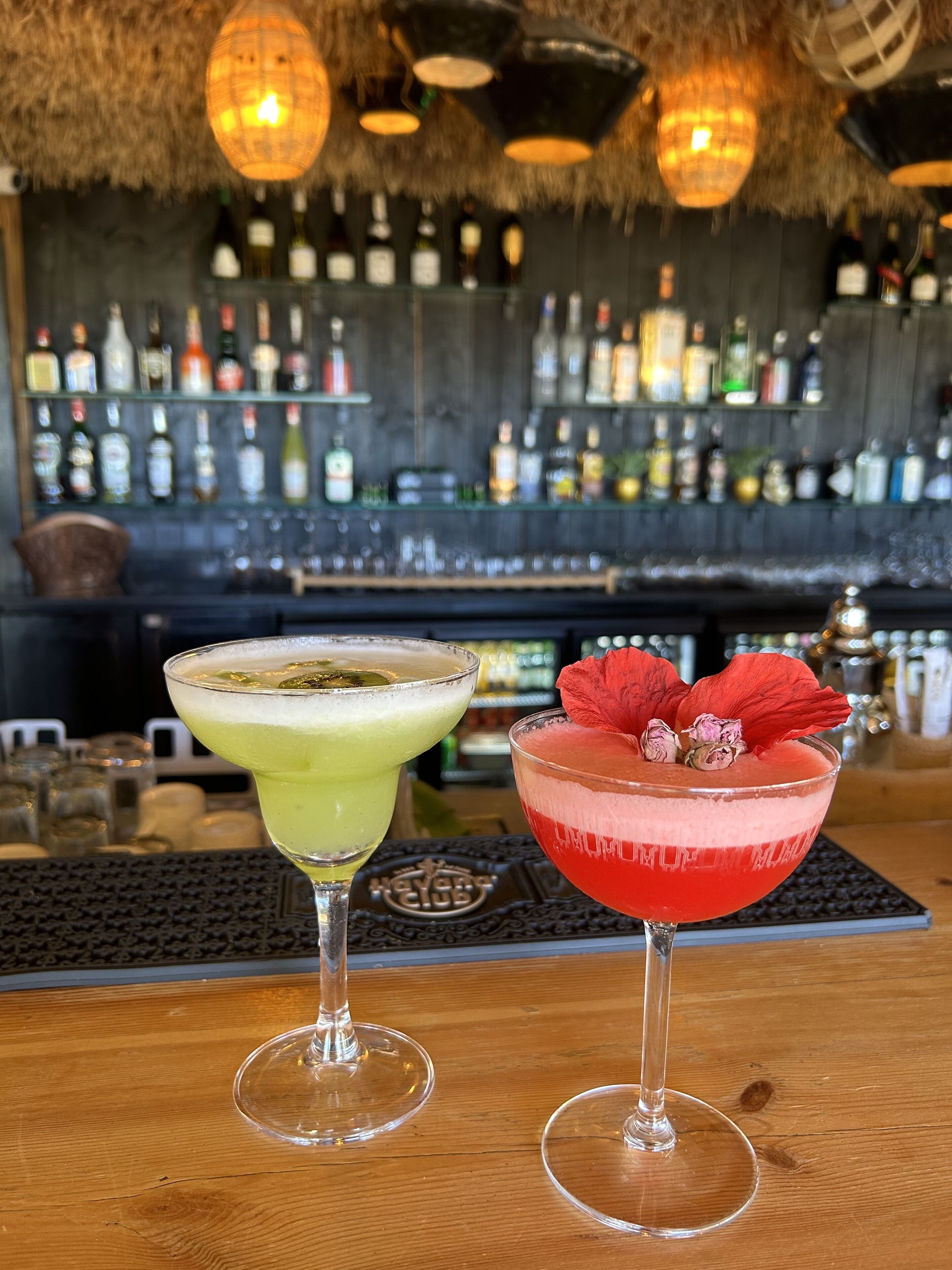 Two cocktails on a bar counter with a backdrop of a bar stocked with various bottles.