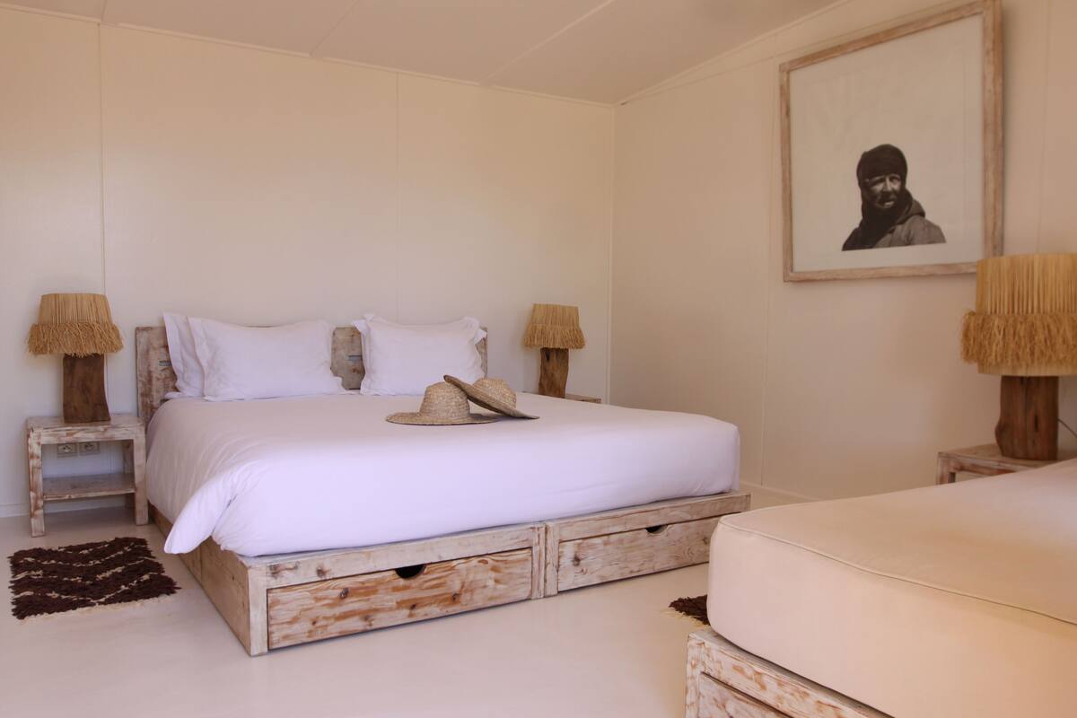 Quarto com uma cama de madeira, duas mesas de cabeceira, um abajur, um quadro na parede e um tapete pequeno.