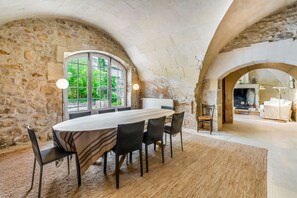Un comedor amplio con una gran mesa y sillas, paredes de piedra y vistas a la vegetación a través de un ventanal.