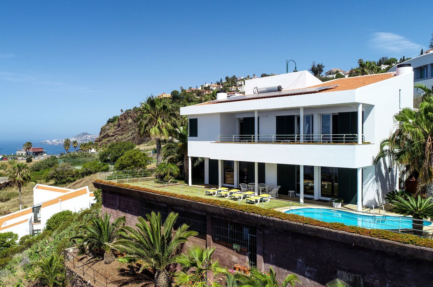 A modern two-story house with a rooftop pool, surrounded by lush greenery and a view of the coastline.