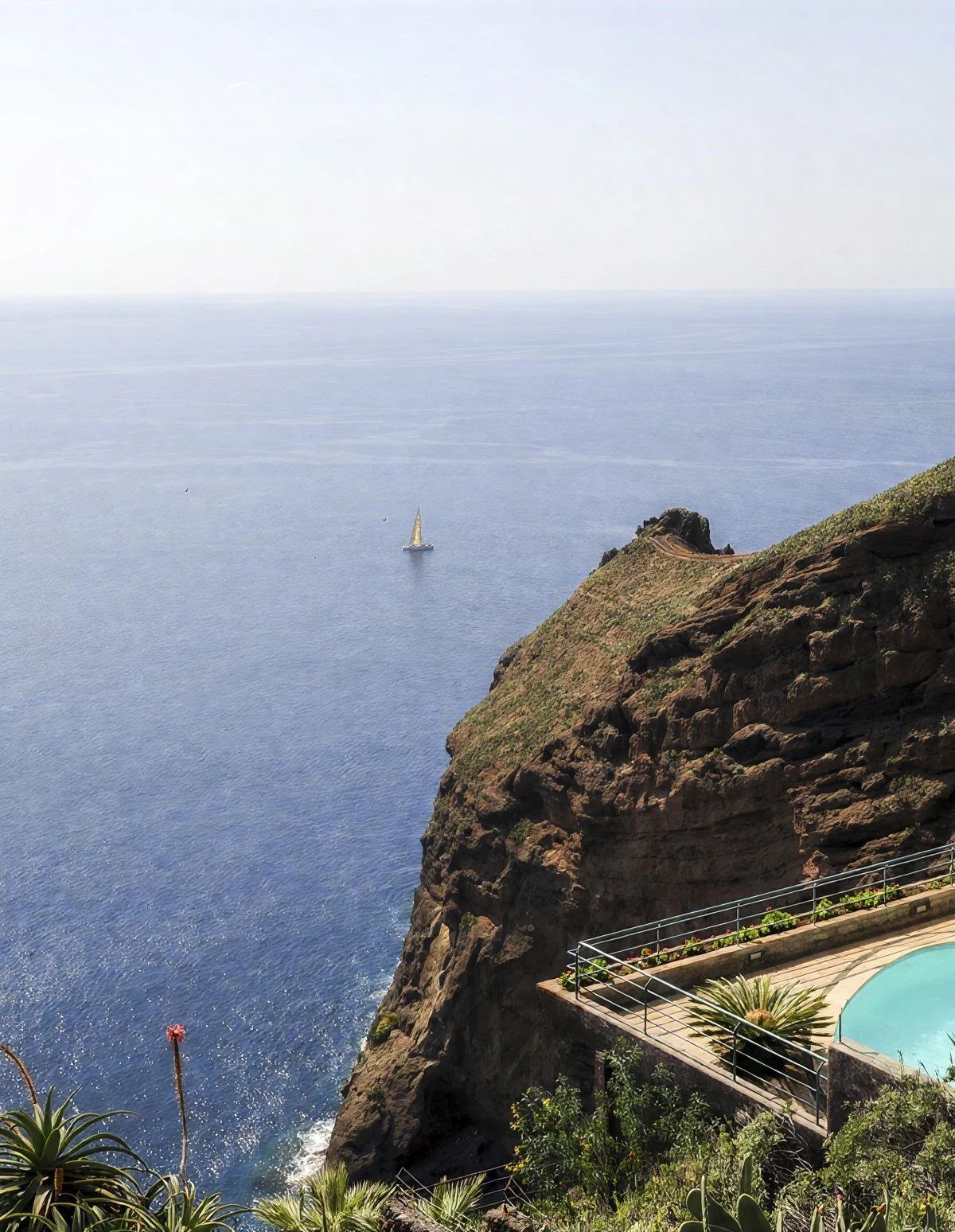 A cliffside view of a coastal area with a pool and a sailboat in the distance.