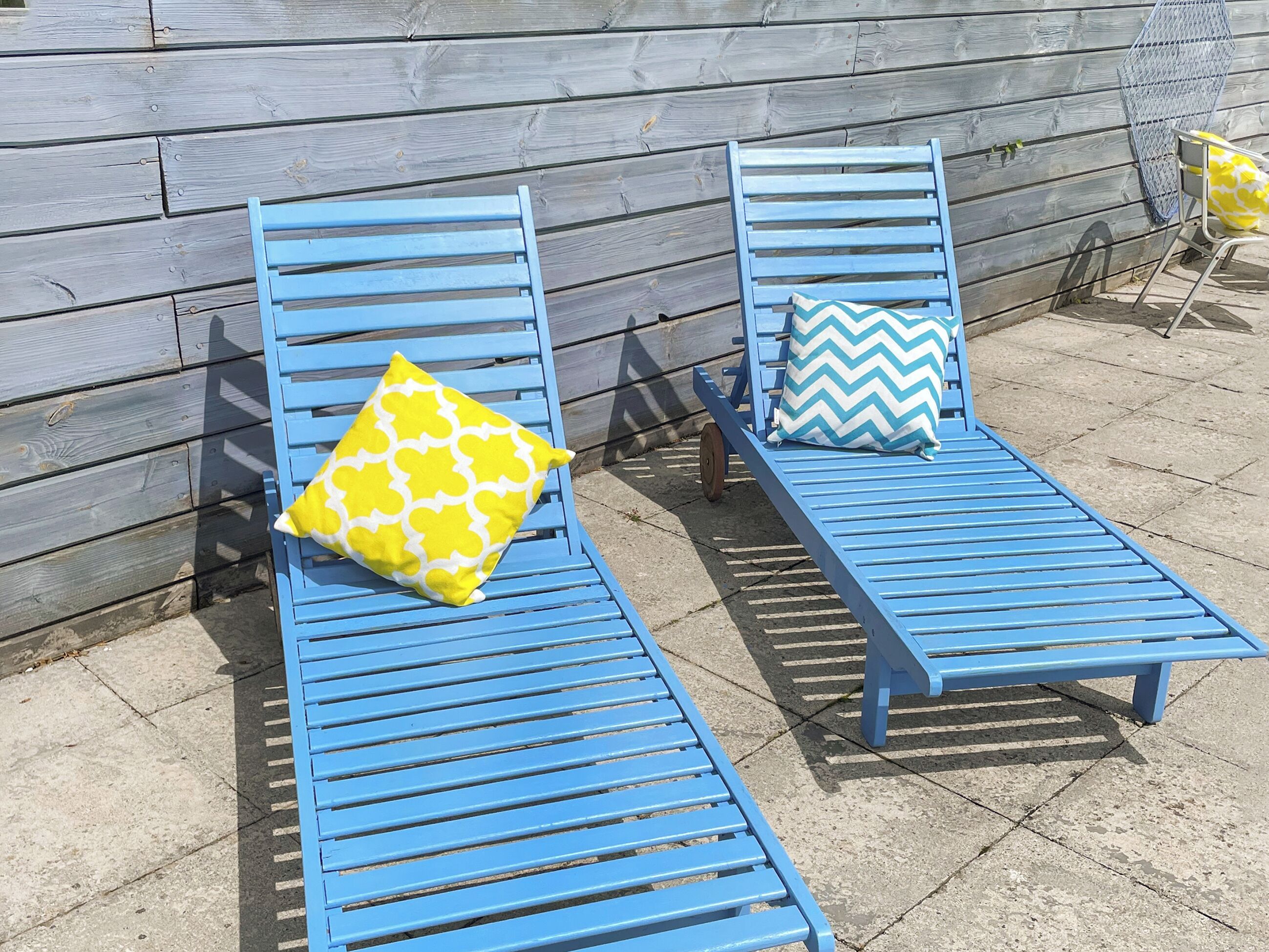 Deux fauteuils de jardin bleus, dotés de coussins à motifs jaunes et bleus, sont placés sur une terrasse en béton, adossés à un mur en bois.