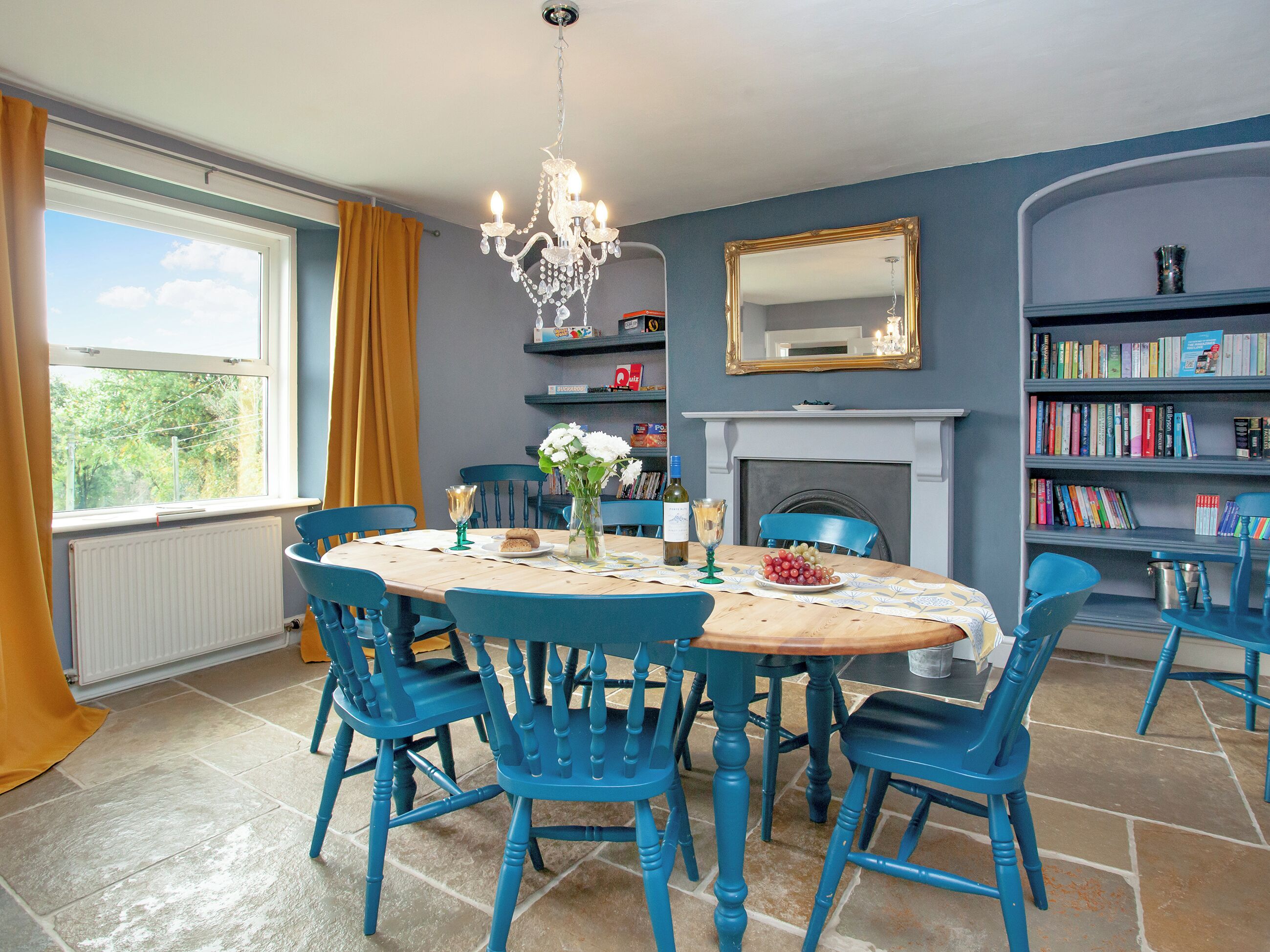 Une salle à manger avec une table dressée pour quatre personnes, des chaises bleues, un lustre, une cheminée et des étagères à livres.