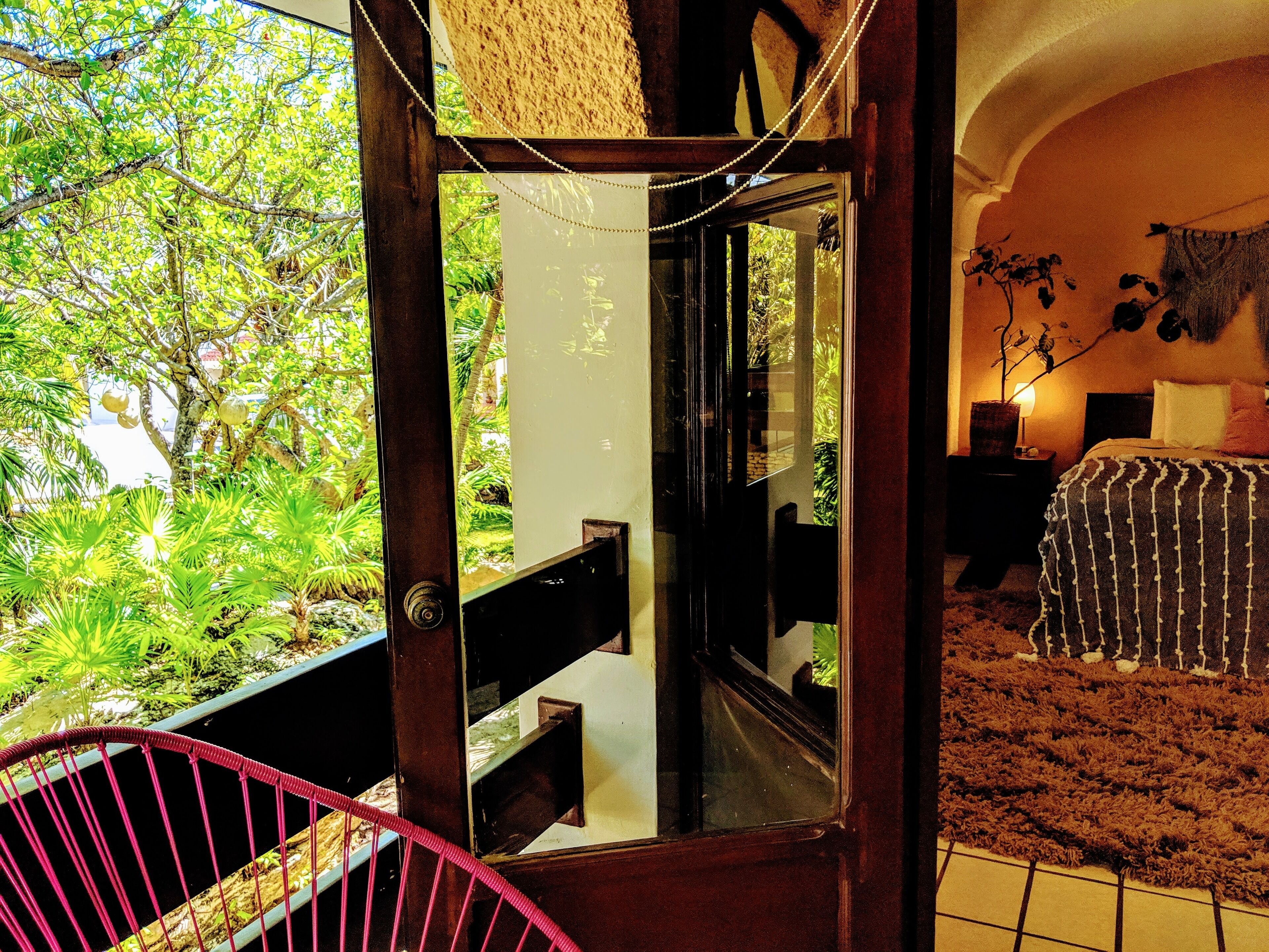 A room with a bed, a nightstand, a potted plant, and a view of greenery outside.