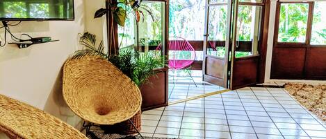 Une pièce avec un téléviseur, un fauteuil en osier, une plante en pot, un fauteuil rose et une vue sur l’extérieur par une porte vitrée.