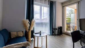 Een woonkamer met een blauwe bank, een kleine ronde tafel met een plant, en uitzicht op een balkon met een tafel en stoelen.