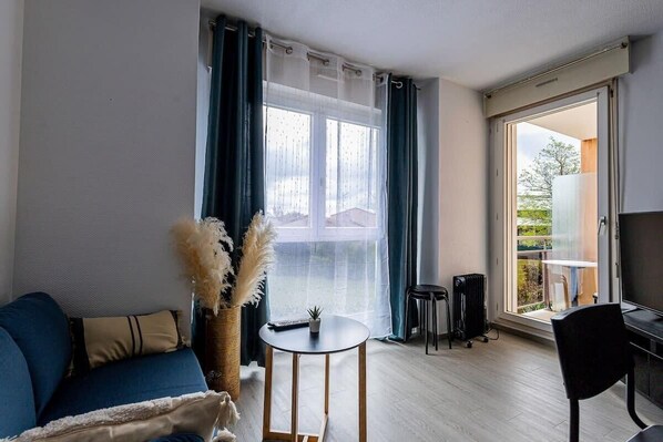 Un salon avec un canapé bleu, une petite table ronde ornée d’une plante, et une vue sur un balcon avec une table et des chaises.